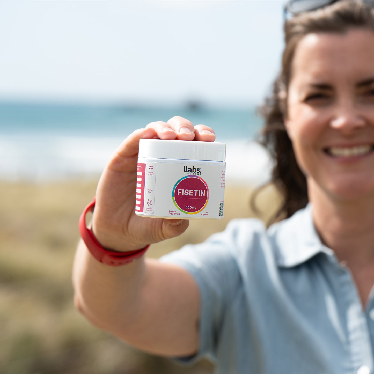 A smiling woman holding up a bottle of llabs Fisetin capsules towards the camera, standing outdoors with a sunny beach background.