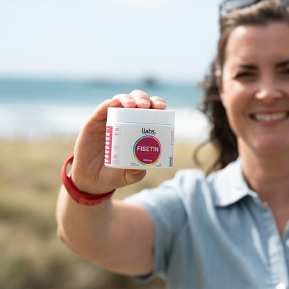 A smiling woman holding up a bottle of llabs Fisetin capsules towards the camera, standing outdoors with a sunny beach background.