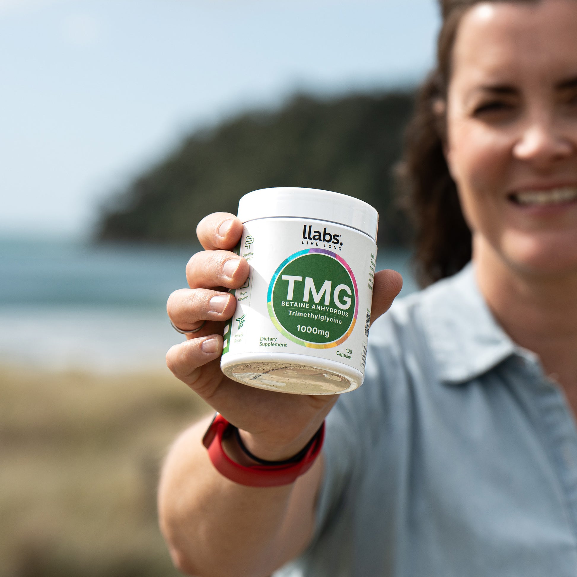 A smiling woman outdoors holding a container of llabs. TMG (Betaine) Capsules, focusing the camera on the product with a natural background to enhance her daily routine.
